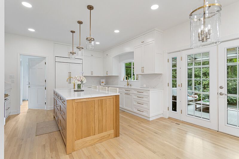nterior design photography in Potomac, Maryland showcasing a bright, modern kitchen with white shaker cabinets, a large natural wood island, gold hardware, and elegant pendant lighting—blending classic design with contemporary charm and outdoor views through French doors.