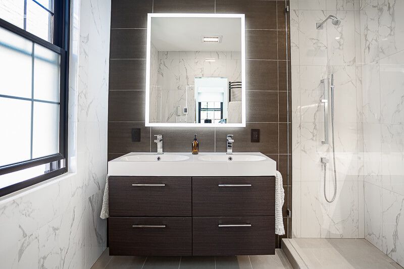 Interior design photography in Chevy Chase, Maryland featuring a sleek modern bathroom with a double vanity, illuminated mirror, dark wood cabinetry, and marble-style tile throughout, creating a refined and contemporary spa-like atmosphere.