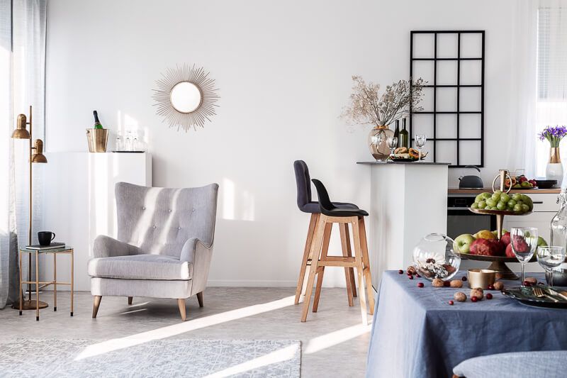 nterior design photography in Washington DC capturing a bright, modern living and dining space with minimalist decor, soft natural light, neutral tones, and elegant furniture including a gray accent chair, bar stools, and a stylish dining table with fresh fruits and glassware.