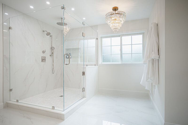 Interior design photography in Washington DC featuring a luxurious all-white bathroom with marble flooring and walls, a spacious glass-enclosed walk-in shower, and an elegant crystal chandelier accentuating the serene, spa-like ambiance.